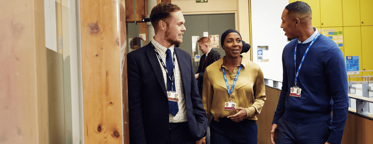 Three teachers walk together and talk in a school corridor, smiling and engaged in conversation.