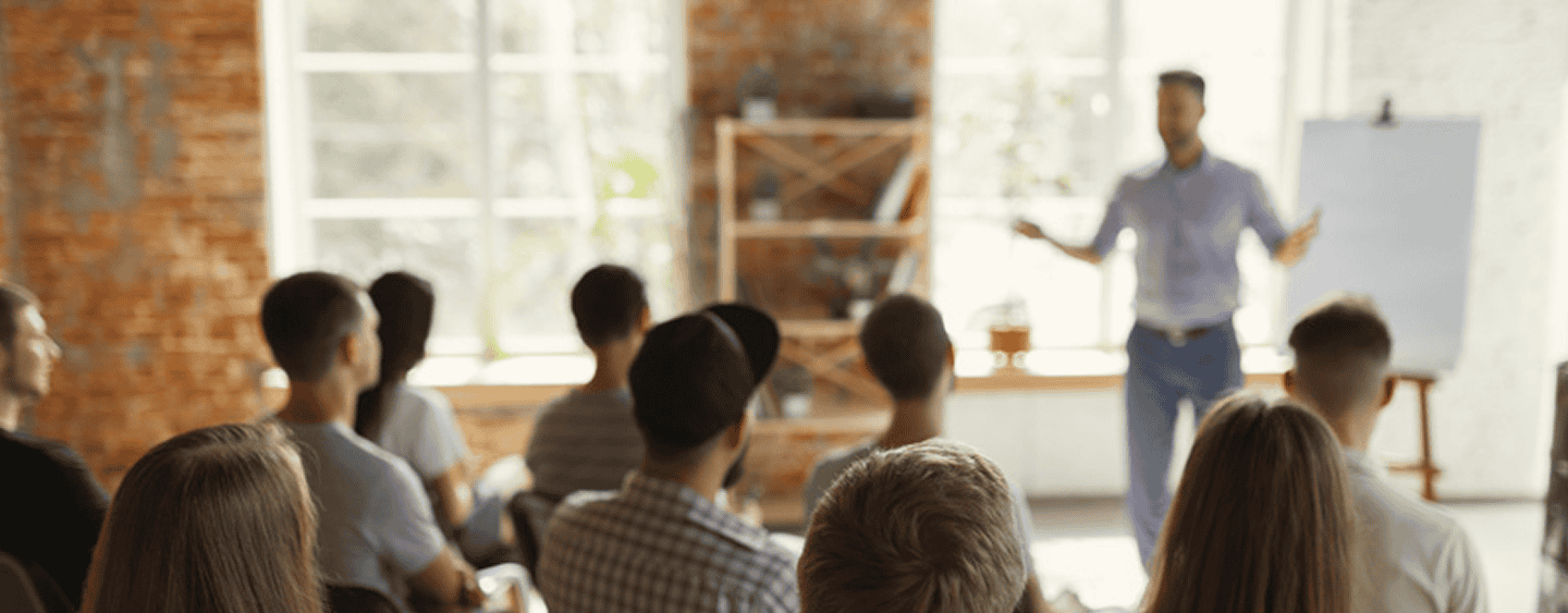 A group of people seated indoors, attentively facing a presenter standing in front of a whiteboard.