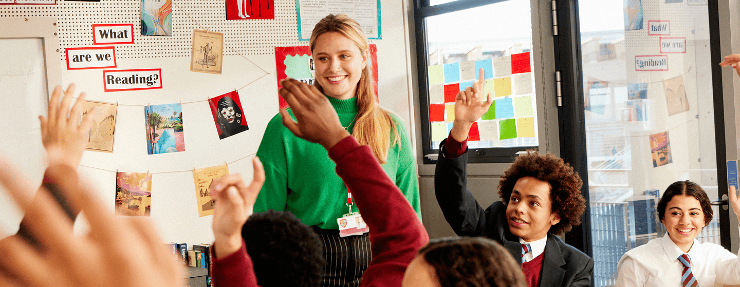 Teacher engaging with enthusiastic secondary school students raising their hands in a classroom decorated with reading materials.